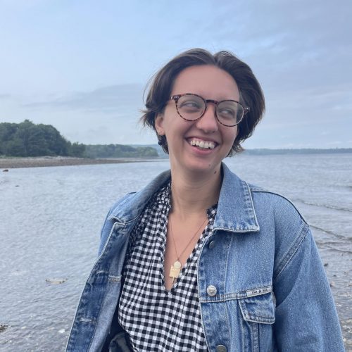 Becca Odell, a white woman with short brown hair and glasses, smiles on the beach