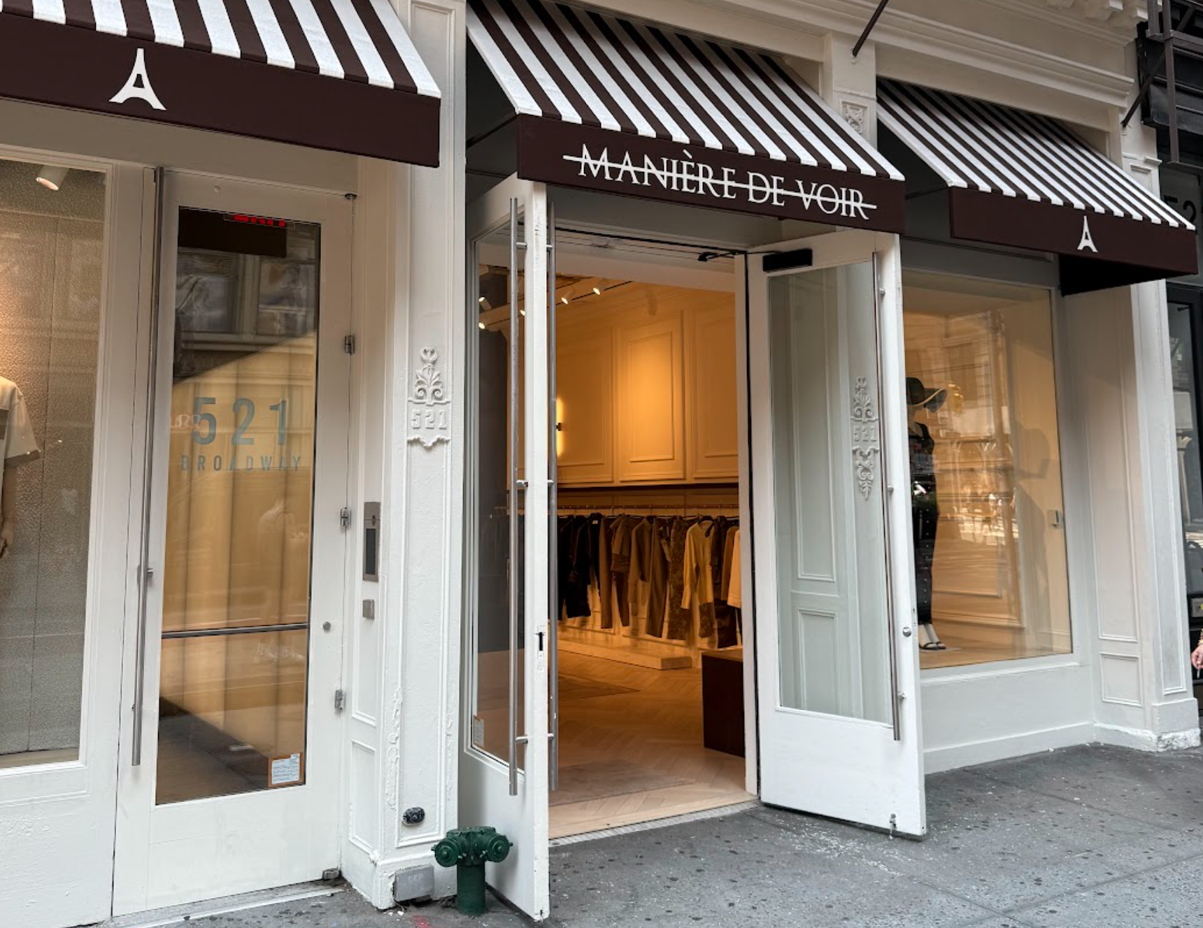 A storefront with two large white doors open to the sidewalk. Inside you can see a warm lit room with women's suits hanging along a far wall. A striped black and white awning shades the entryway.