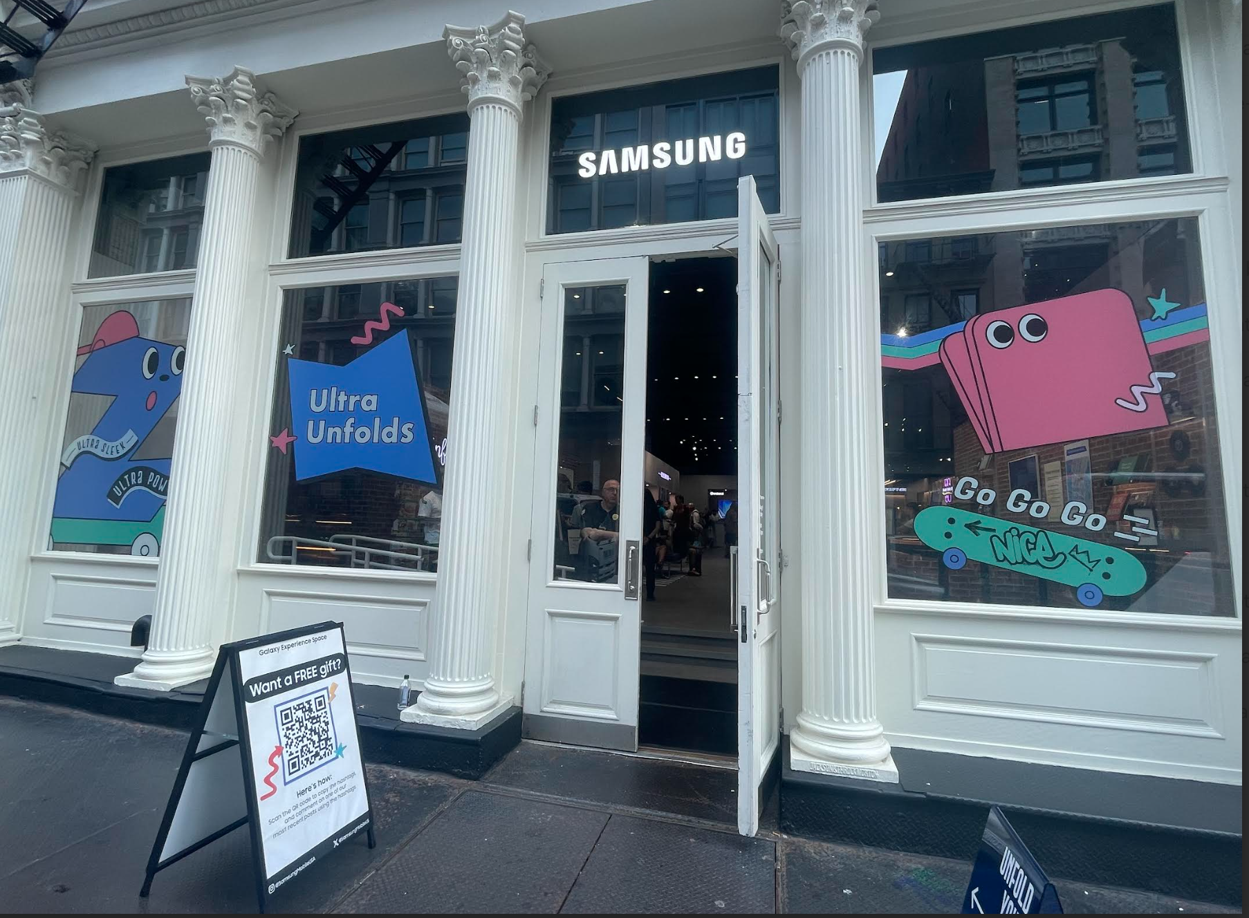This storefront features four windows with cartoon drawings of Samsung phones and the phrases "Ultra Unfolds" and "Go Go Go". Above the open door is a lit up sign for Samsung.  