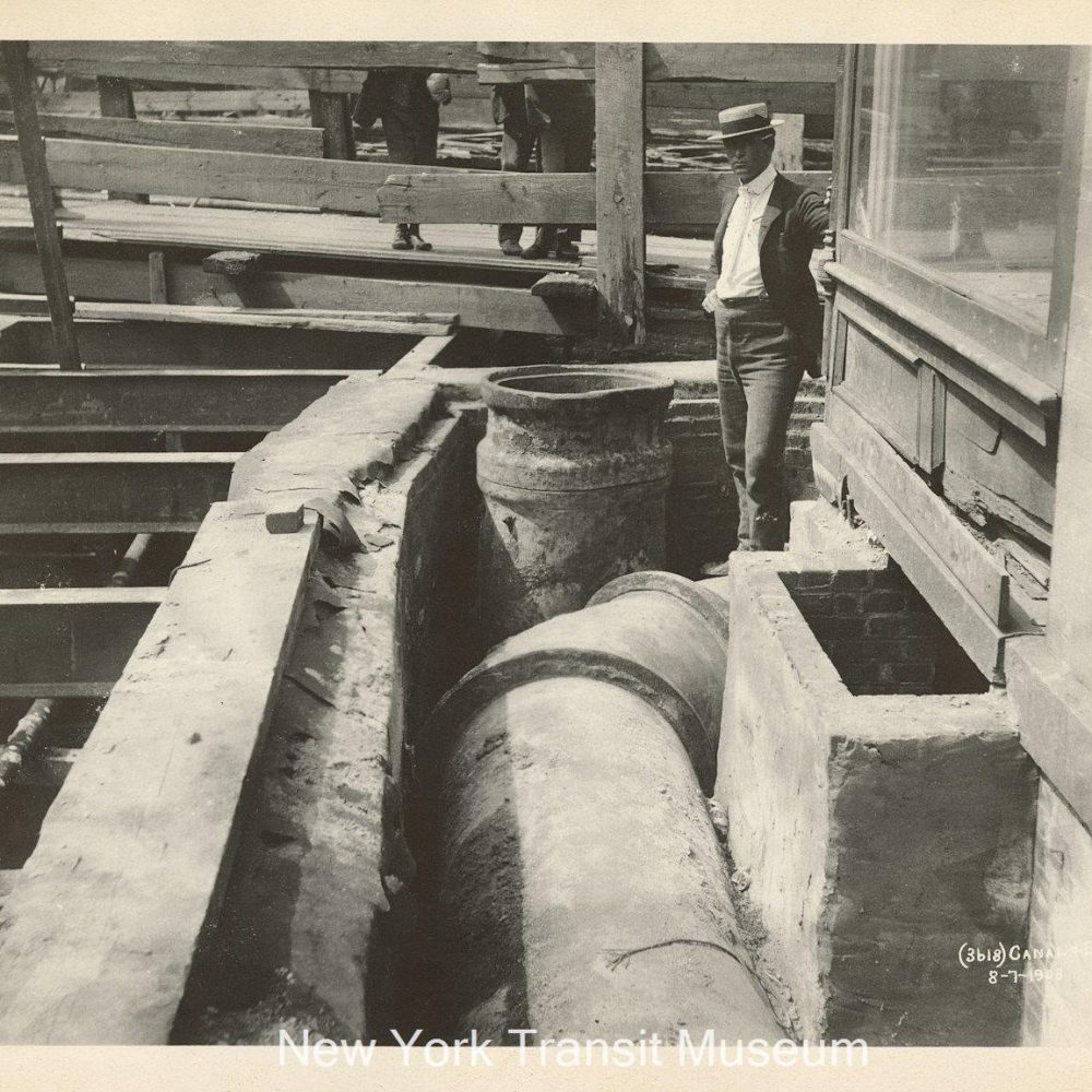A black and white photo showing Canal St excavated for subway construction. The street has been torn up to reveal sewer pipes, and wooden boards have been placed to bridge the gap. A man stands near the open street.