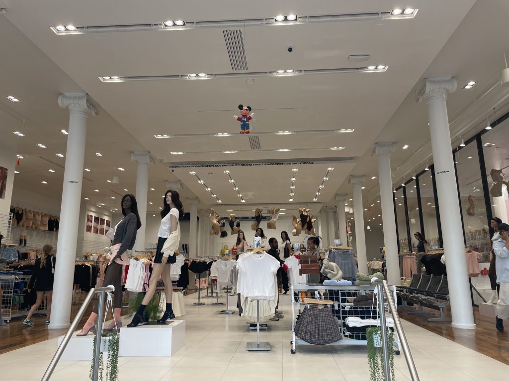 Mannequins and clothing racks in a large room with white marble floors, overhead lighting, and decorative columns. A Mickey Mouse stuffed animal hangs from the ceiling. 