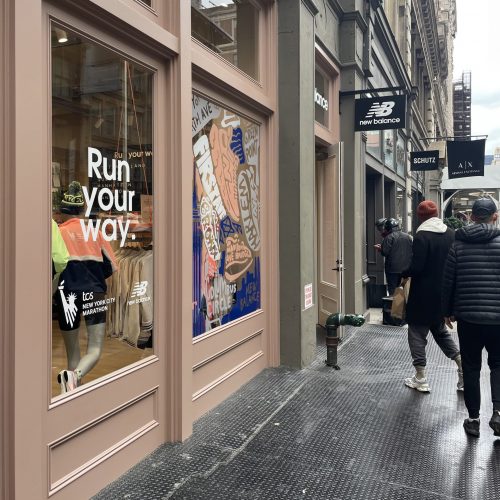 Glass windows of a storefront on a busy street say "Run your way". The black New Balance sign extends out from the building over the heads of pedestrians