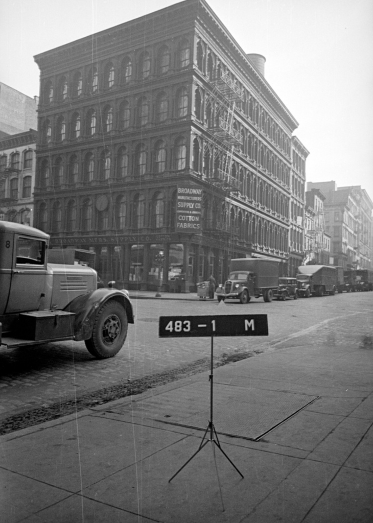 A black and white image shows SoHo in the 40s, with a line of old fashioned box trucks parked on the road and a large, ornate cast iron building on the corner.