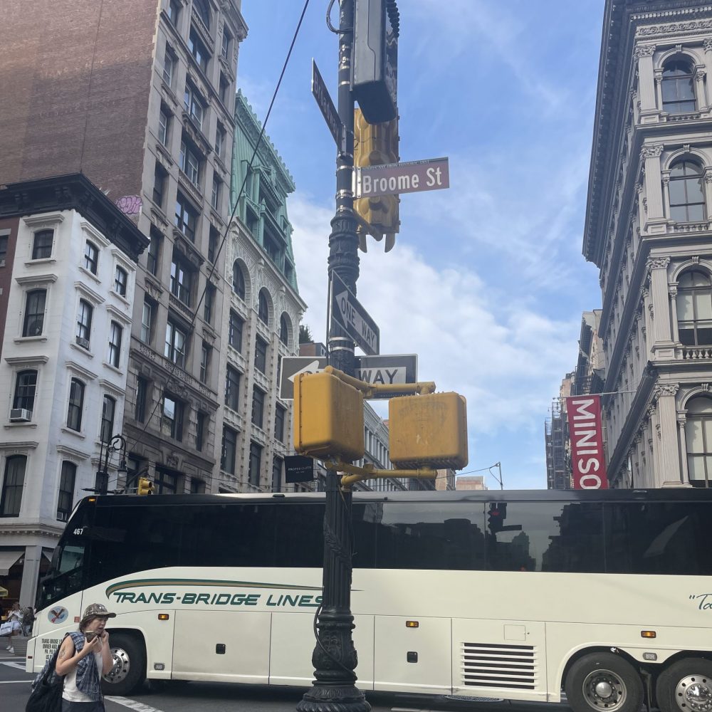 Image shows a bus and pedestrians crossing Broome St on a sunny day