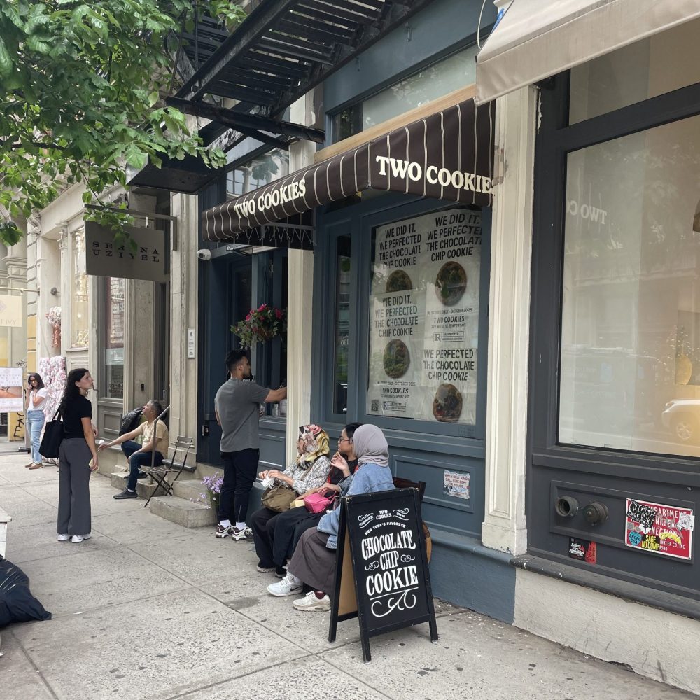 A black and white awning over a sidewalk reads "Two Cookies". Two people wait to order at the window and three people sit on a bench, enjoying cookies.