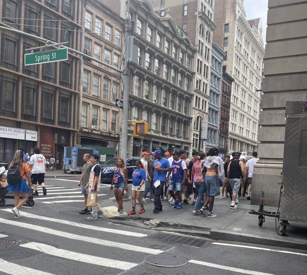 A crowd gathers at an intersection, many sporting orange and blue Knicks garb. It is an overcast day in SoHo, and a streetsign shows they are at Spring St.